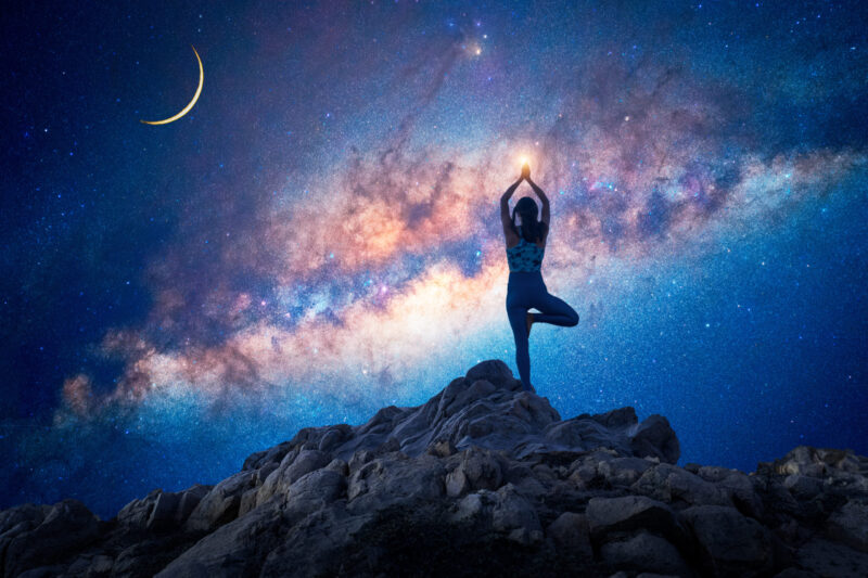 silhouette of a woman standing in yoga tree position on the rock or mountain with Milky Way background